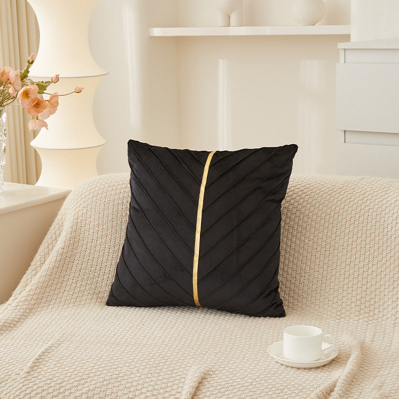 A black square throw pillow with a central gold stripe and diagonal stitched texture, resting on a cream-colored textured sofa in a bright, modern living room setting.