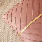 A close-up of a dusty pink velvet throw pillow with vertical channel tufting and a diagonal gold metallic trim, resting on a textured beige woven rug.