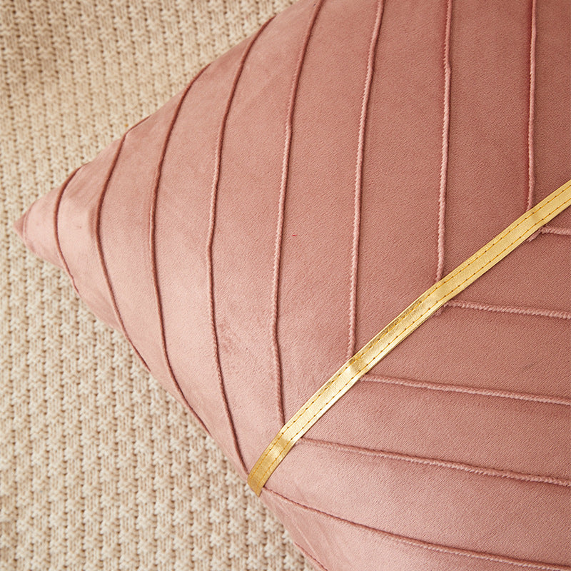 A close-up of a dusty pink velvet throw pillow with vertical channel tufting and a diagonal gold metallic trim, resting on a textured beige woven rug.