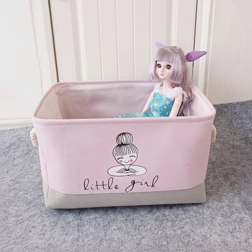 Pink storage basket with 'little girl' design containing a doll on a gray surface.