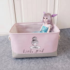 Pink storage basket with 'little girl' design containing a doll on a gray surface.