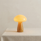 A mushroom-shaped table lamp with a glowing warm yellow cap and a brownish-yellow stem, displayed on a light beige fabric-covered surface against a neutral beige background.