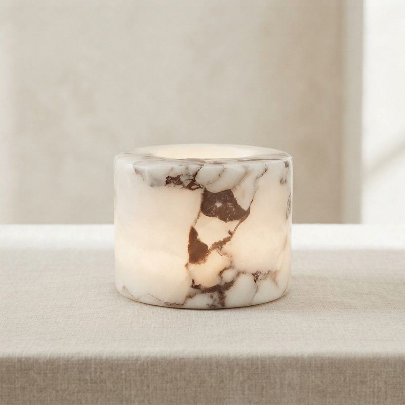 A small cylindrical marble candle holder or candle base with a warm white and brown veined pattern, glowing softly from within, set on a neutral beige fabric surface.