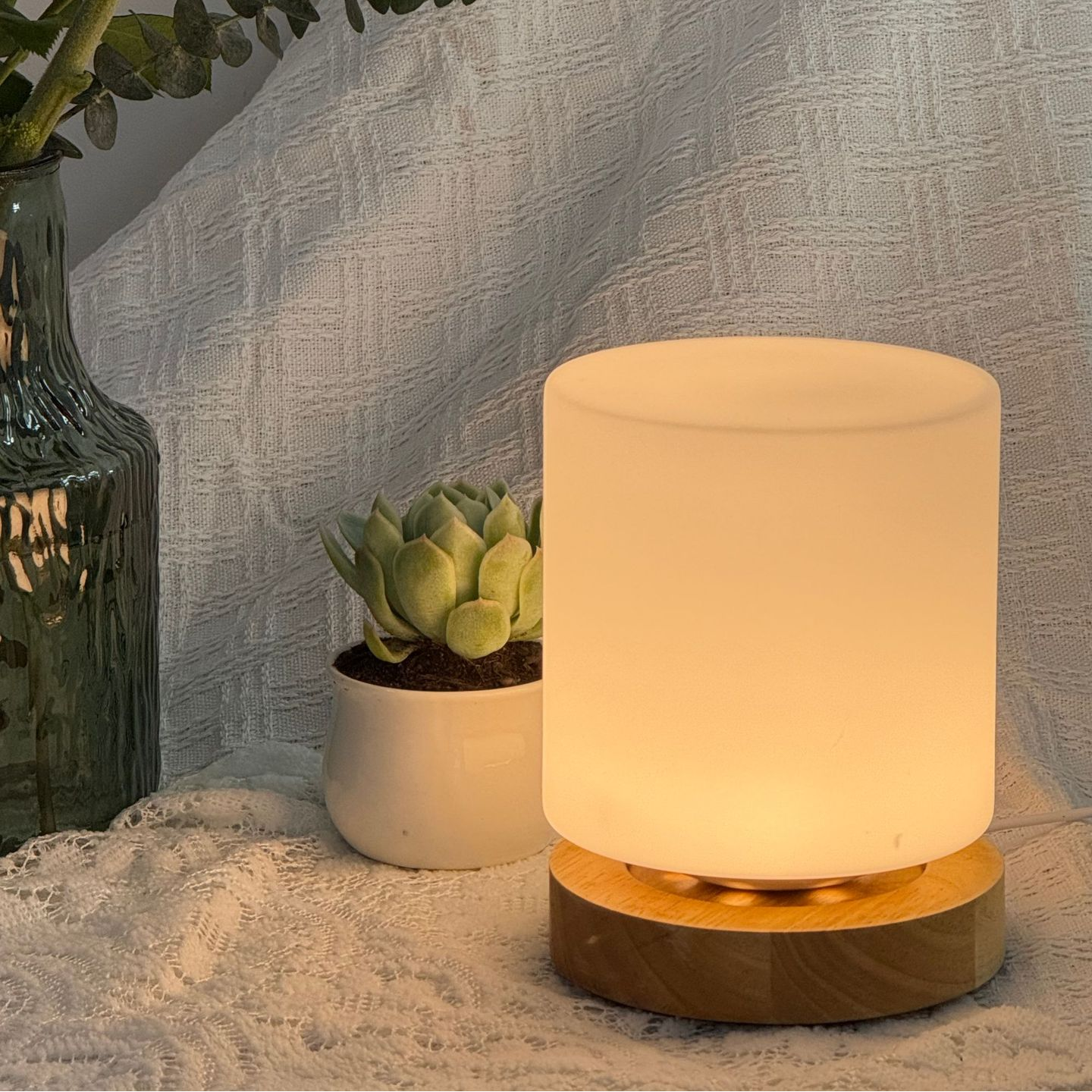 Frosted glass lamp with wooden base on a textured surface with a plant and decorative vase.