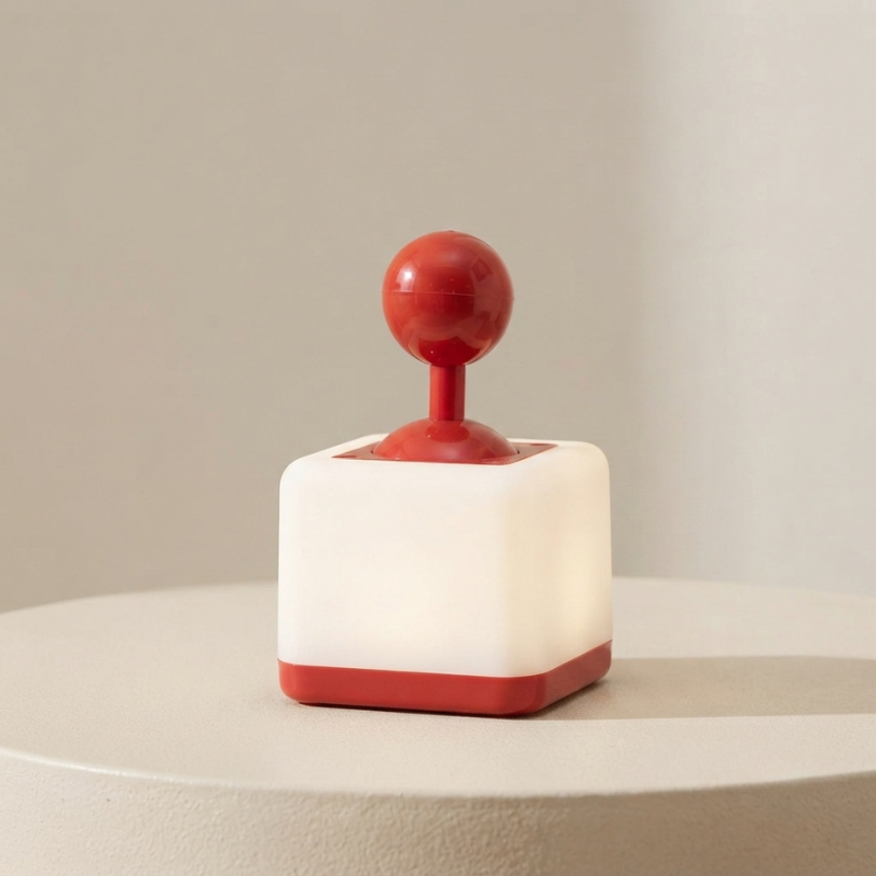 A small modern lamp shaped like a red joystick with a glowing white square base, shown on a round pedestal against a soft beige background.