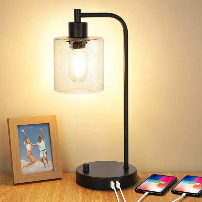 Black table lamp with a glass shade on a wooden surface, next to two smartphones charging.
