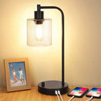 Black table lamp with a glass shade on a wooden surface, next to two smartphones charging.