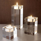 Set of three square glass candle holders with candles on a reflective surface.