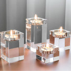 Four clear acrylic square candle holders with lit tealight candles, arranged on a reflective metallic surface against a soft gray background.