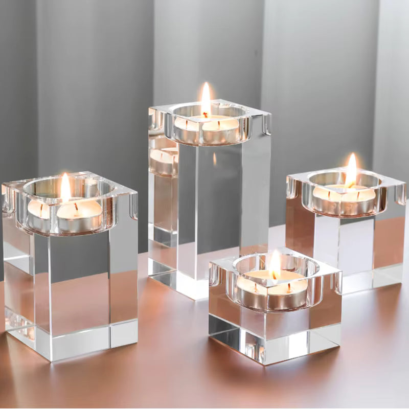 Four clear acrylic square candle holders with lit tealight candles, arranged on a reflective metallic surface against a soft gray background.