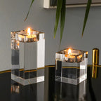 Two clear acrylic square candle holders with metallic silver rims, each holding a lit tealight candle, displayed on a reflective black surface against a gray wall with green leaves hanging in the foreground.