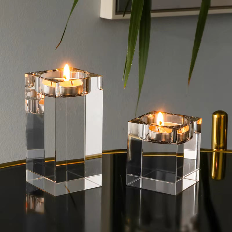Two clear acrylic square candle holders with metallic silver rims, each holding a lit tealight candle, displayed on a reflective black surface against a gray wall with green leaves hanging in the foreground.