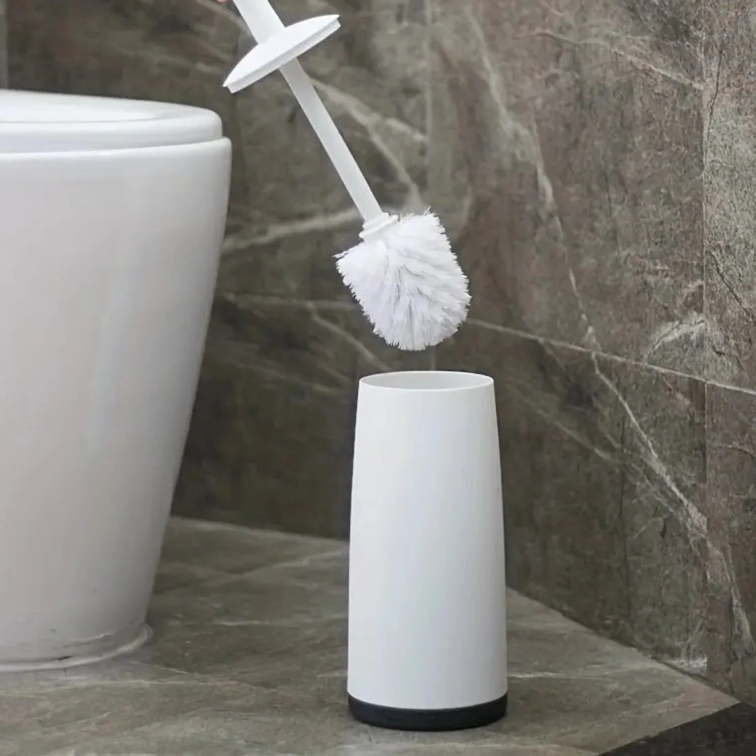 White toilet brush with black base in a holder next to a white toilet on a tiled bathroom floor.