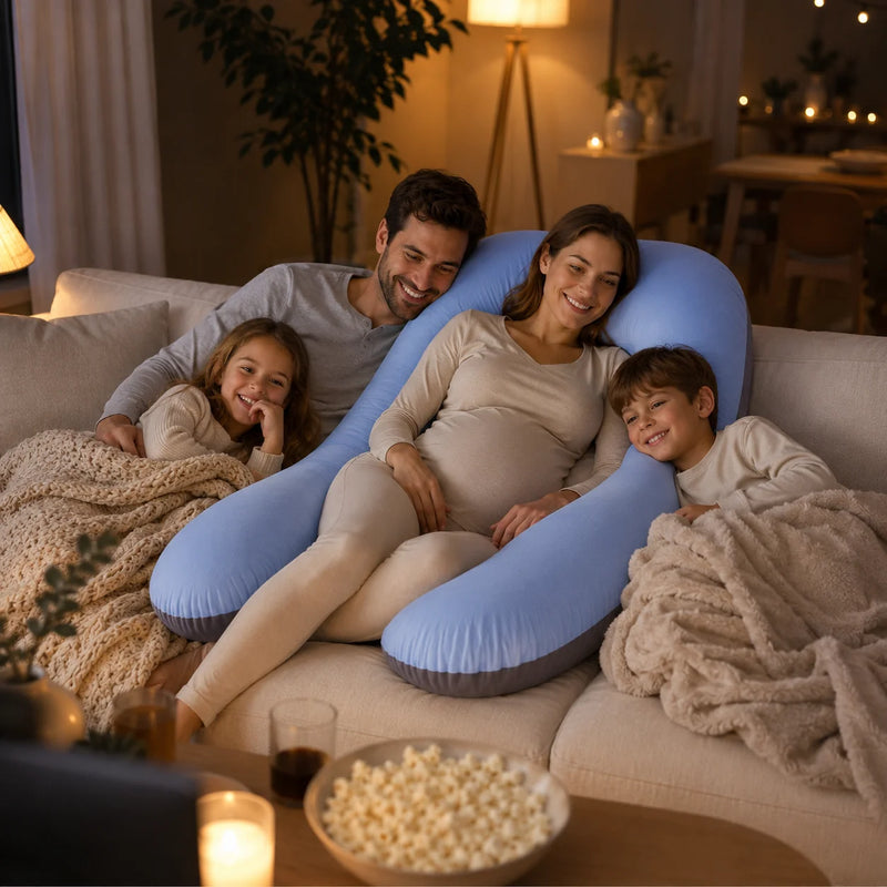 A pregnant woman and her three children relax on a beige sofa in a warmly lit living room, using a large light blue U-shaped pregnancy support pillow. A bowl of popcorn and a laptop are visible in the foreground.