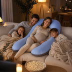A pregnant woman and her three children relax on a beige sofa in a warmly lit living room, using a large light blue U-shaped pregnancy support pillow. A bowl of popcorn and a laptop are visible in the foreground.