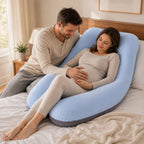 A pregnant woman reclines on a bed using a light blue U-shaped pregnancy support pillow, with a man sitting beside her on the bed.