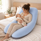 A woman in a bedroom sits on a bed using a large U-shaped baby support pillow to hold and support a baby. The pillow is light blue with a dark gray base, and the room has a wooden headboard, white bedding, and a bedside lamp.
