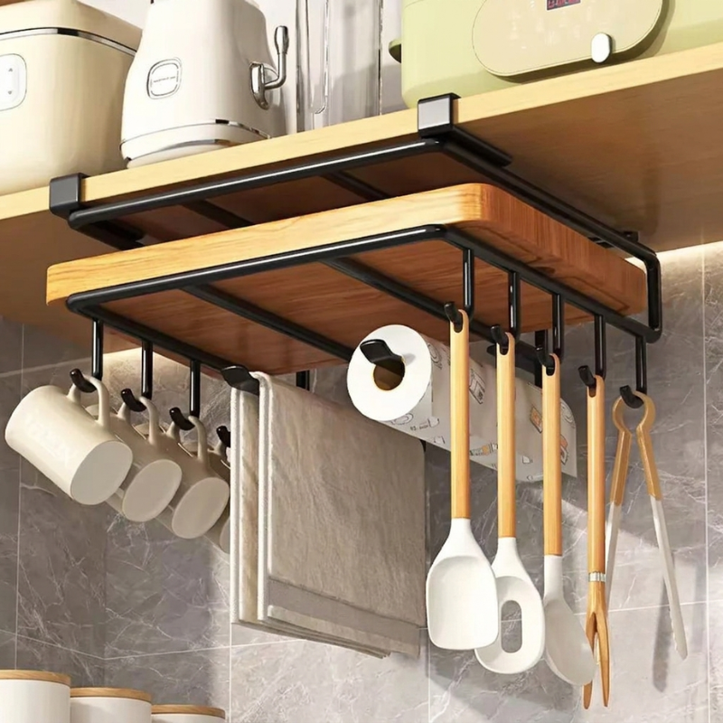 A wall-mounted kitchen organizer rack with a wooden board on a black metal frame, shown installed under a shelf in a kitchen. Hanging utensils include white spatulas, wooden spoons, tongs, a towel, and a roll of paper towels, with white mugs hanging on the left.