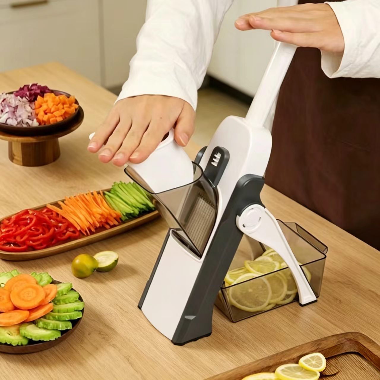 A white and dark gray manual mandoline slicer being operated by a person on a wooden kitchen counter, with sliced vegetables and lemon slices nearby.