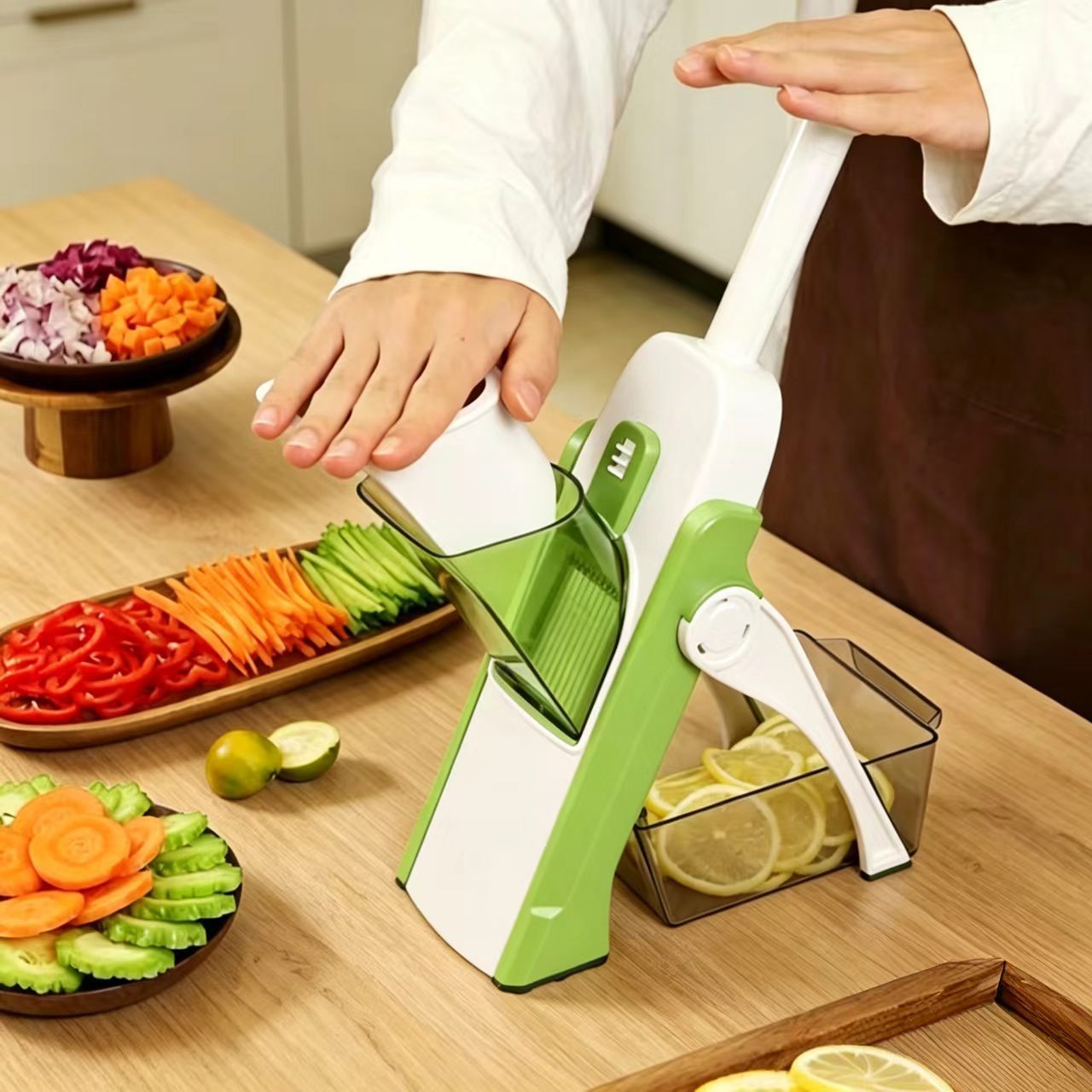 A green and white manual mandoline slicer being demonstrated on a wooden kitchen counter, with a person's hands visible operating the device. Sliced vegetables including carrots, cucumber, red pepper, and lemon slices are arranged nearby.
