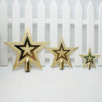 Three gold metallic star-shaped Christmas tree toppers in different sizes, arranged from largest to smallest against a white picket fence background.