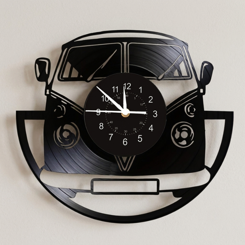 A black metal wall clock shaped like a classic Volkswagen bus, made from cut vinyl records, with a round black clock face and white hands, mounted on a light beige wall.