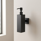 A matte black rectangular soap dispenser with a matching wall-mounted pump and backplate, mounted on a warm beige wall next to a window.