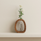 A small oval wooden vase with a clear glass tube center holding a single white flower with green leaves, displayed on a white shelf against a warm beige wall.