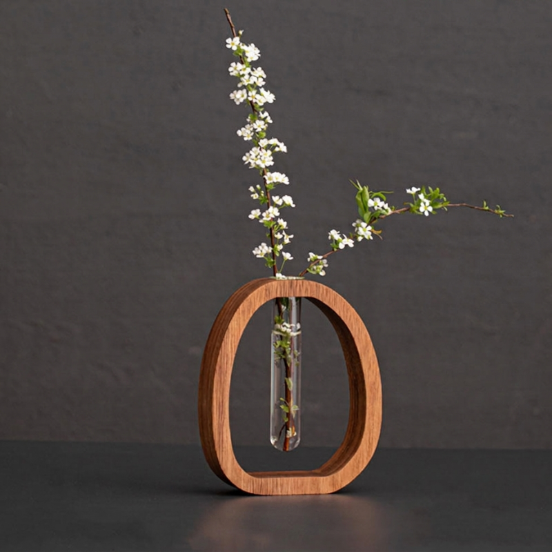 A minimalist wooden oval vase with a clear glass vial holding a flowering branch with small white blossoms, set against a dark gray textured background.
