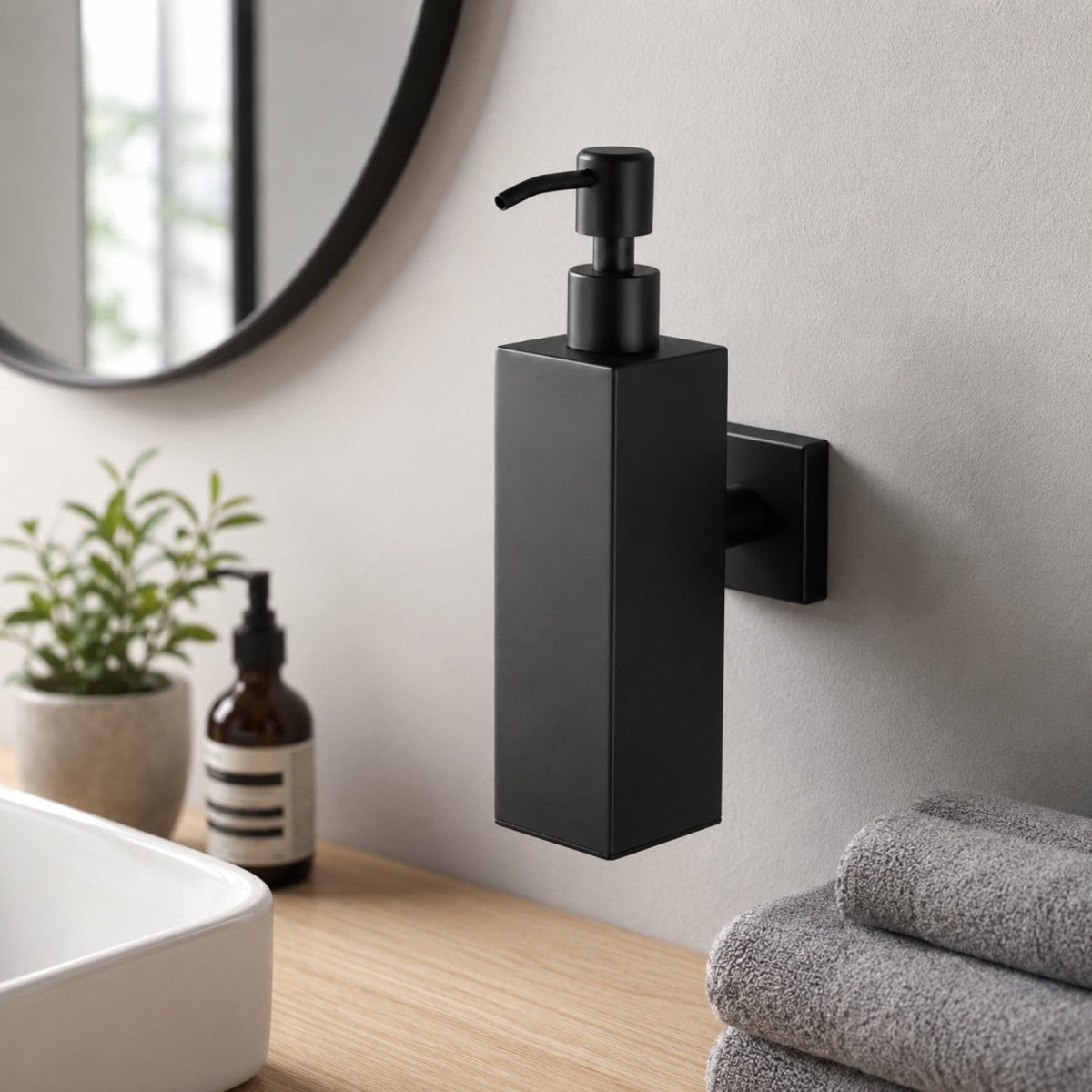 Matte black wall-mounted soap dispenser with a rectangular body and pump, shown installed on a bathroom wall above a sink with folded towels and a small plant nearby.