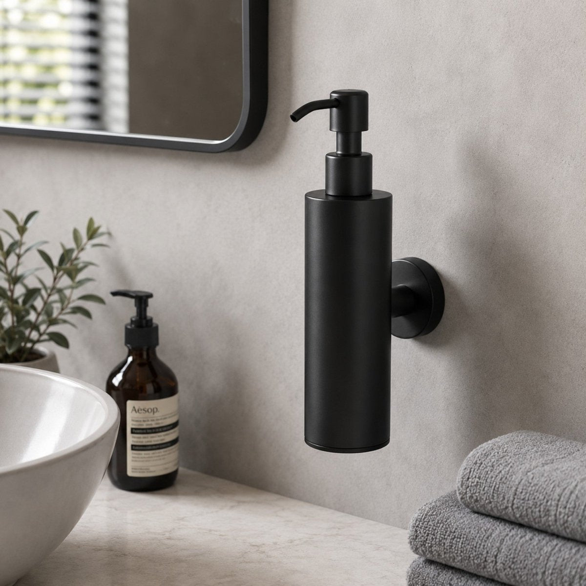 A matte black wall-mounted pump dispenser bottle with a cylindrical body and a curved top spout, shown installed on a bathroom wall beside a sink, soap bottle, plant, and folded towels.