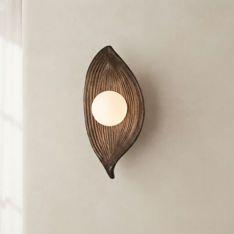 A leaf-shaped wall sconce with a dark brown ribbed frame and a glowing warm white circular light, mounted on a plain beige wall.
