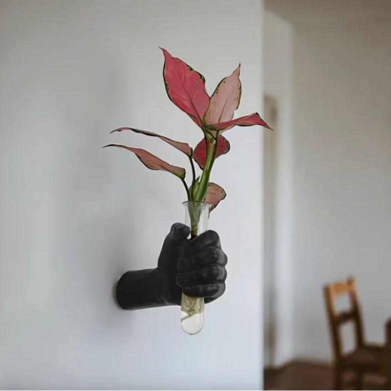 Hand wearing a black glove holding a small plant in a clear vase against a plain background