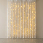 Warm white curtain string lights glowing through a white sheer curtain panel hanging in front of a neutral beige wall.