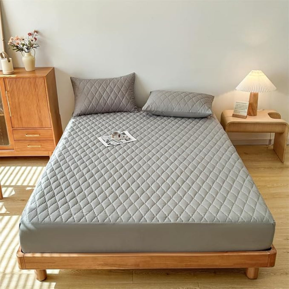 Gray quilted mattress cover on a bed in a bedroom setting with wooden furniture.