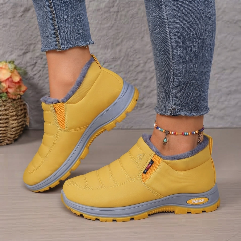 A pair of women's casual slip-on shoes in mustard yellow with gray soles and a gray fuzzy lining, shown on a model wearing blue jeans.