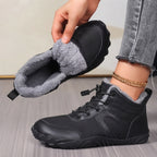 A person holds up a black winter boot with a gray fleece lining, while wearing the matching boot. The boot has a black synthetic upper, lace-up front, and thick gray fleece collar.