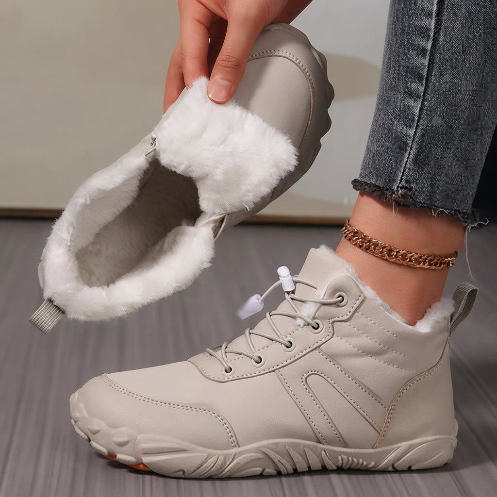 A person wearing a light beige winter lace-up boot with white faux fur trim, while holding the second boot to show the interior. The person is wearing gray jeans and a gold chain anklet, standing on a gray wood floor.