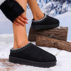 Black slip-on winter slippers with white zigzag stitching around the collar, shown on a model's foot and other foot, set in a snowy outdoor scene with logs.