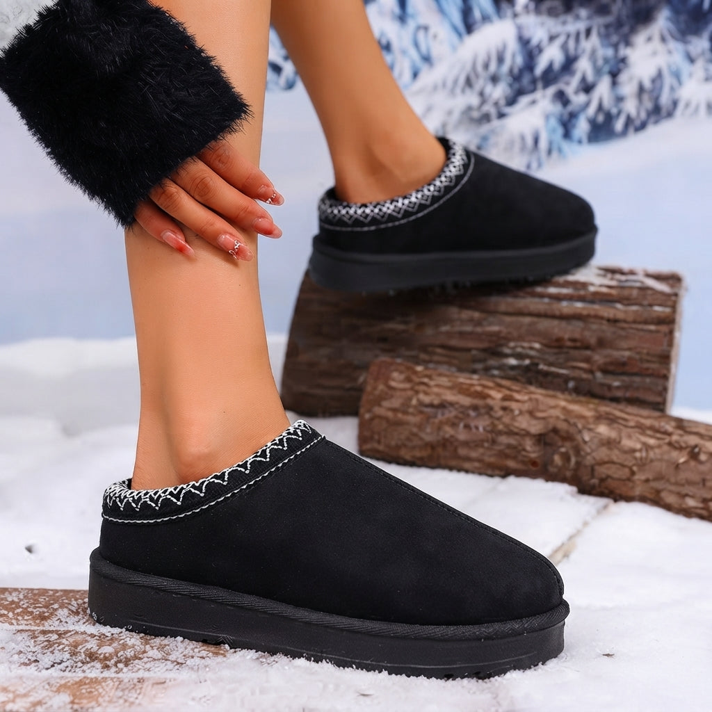 Black slip-on winter slippers with white zigzag stitching around the collar, shown on a model's foot and other foot, set in a snowy outdoor scene with logs.