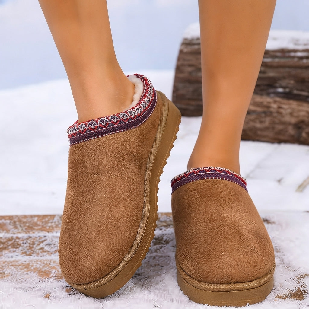 A person wearing brown suede-like slip-on slippers with a multicolor patterned trim, standing outdoors in a snowy setting.