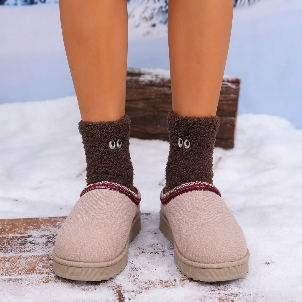 A person standing outdoors in snow wearing light beige slip-on slippers with a dark brown fuzzy sock featuring cartoon eyes, plus a rustic wooden bench in the background.