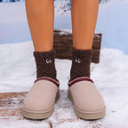 A person standing outdoors in snow wearing light beige slip-on slippers with a dark brown fuzzy sock featuring cartoon eyes, plus a rustic wooden bench in the background.