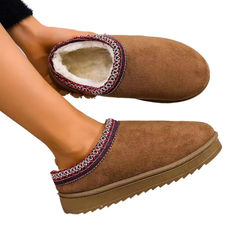 A pair of women's slip-on slipper shoes in brown suede, featuring a cream sherpa lining and a decorative red, white, and blue geometric trim around the collar. One shoe is shown being worn on a foot, while the other is held above it against a solid white background.