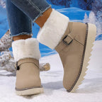 A pair of beige faux-suede ankle winter boots with fluffy white faux-fur trim and side buckle details, shown on a model's feet in blue jeans against a snowy outdoor backdrop.