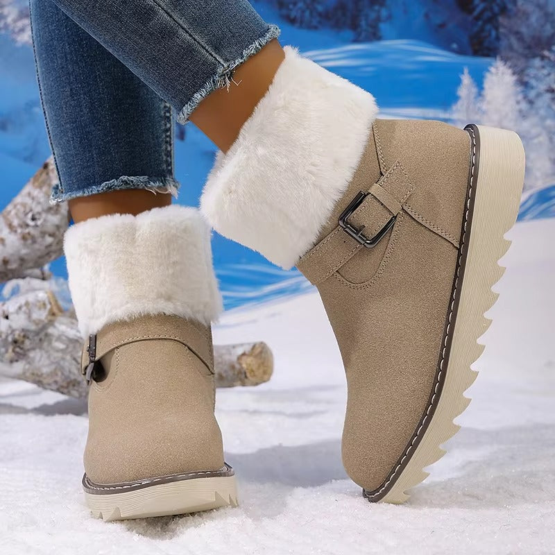 A pair of beige faux-suede ankle winter boots with fluffy white faux-fur trim and side buckle details, shown on a model's feet in blue jeans against a snowy outdoor backdrop.