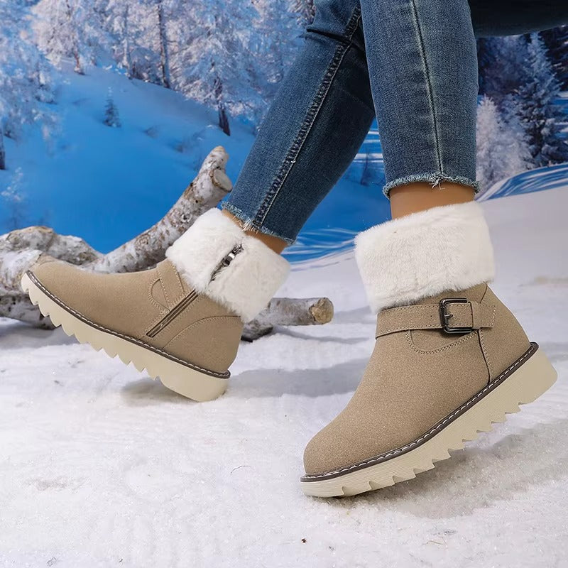 A person wearing beige ankle-height winter boots with white faux-fur cuffs and side zippers, standing in a snowy outdoor-style setting with a blue winter forest backdrop.