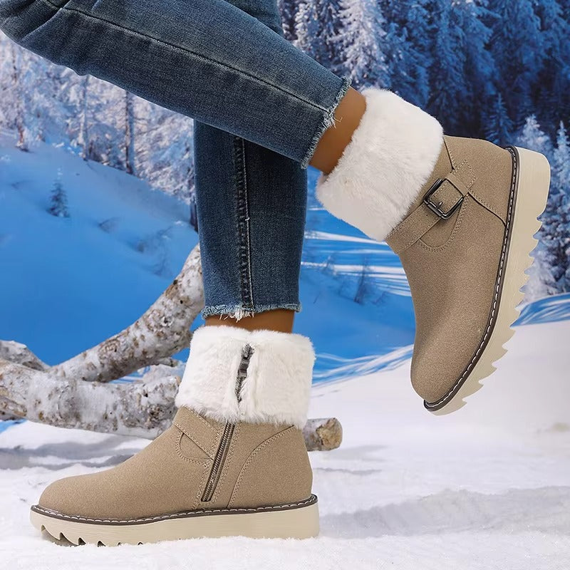 A person wearing beige ankle-height winter boots with white faux-fur trim and side zippers, paired with blue jeans, posed against a snowy outdoor scene with trees and logs.