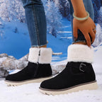 A person wearing black ankle-height winter boots with thick white faux-fur trim and a buckle strap, standing in a snowy outdoor scene with a winter forest backdrop.
