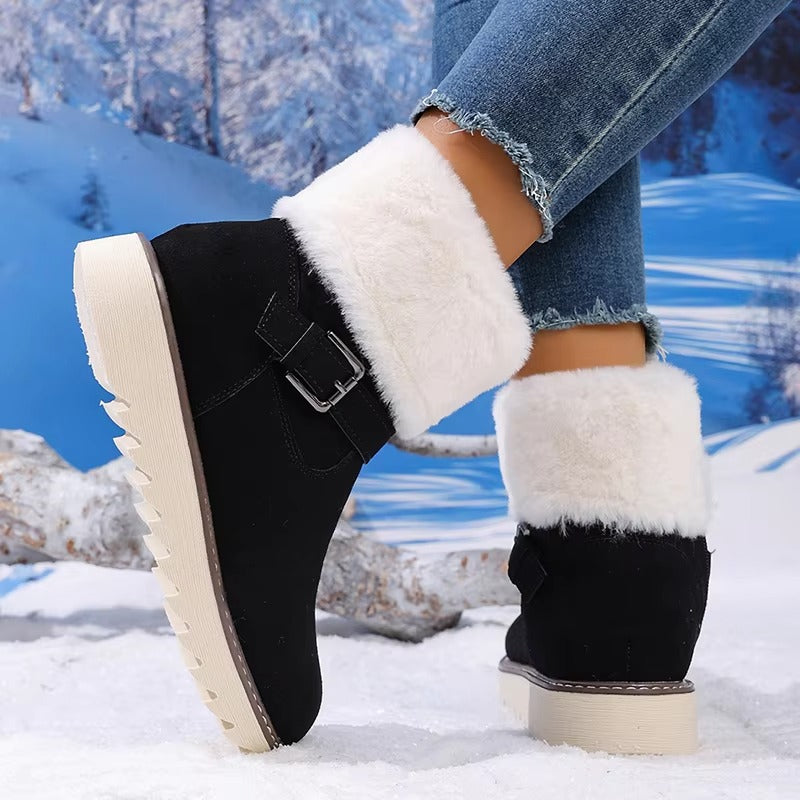 A person wearing black ankle winter boots with fluffy white faux-fur cuffs and a side buckle strap, standing in snow with a snowy forest backdrop.